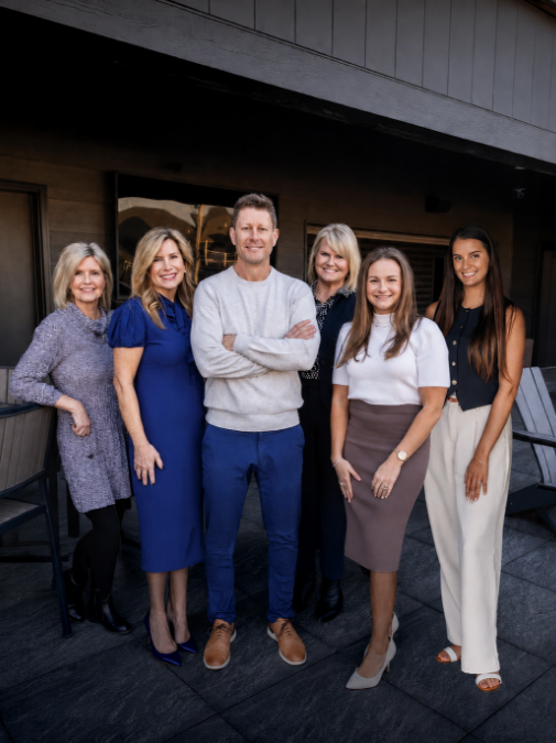 Team of six financial professionals standing together outdoors in front of their office, smiling and dressed in professional business attire.