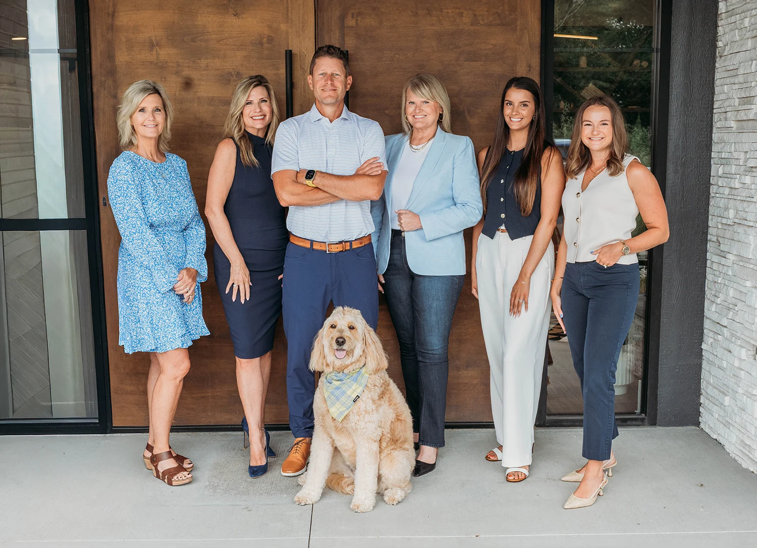 Group portrait of six adults standing with a dog in front of a wooden entrance